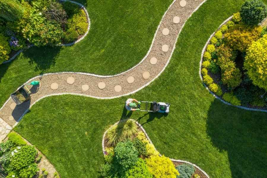 Vue aérienne d'un jardin paysager avec une allée sinueuse et un jardinier tondant la pelouse.